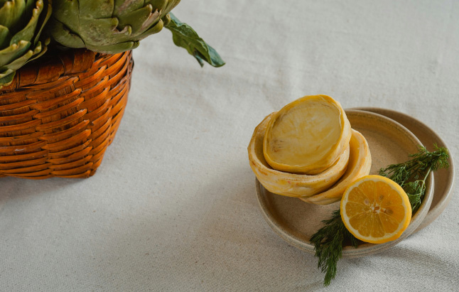 A woven basket filled with artichokes alongside a white plate with lemon and leaves.